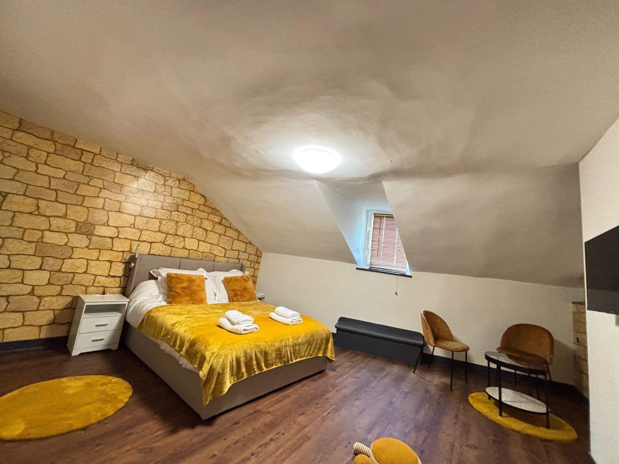 Een slaapkamer met een stenen muur, een bed met een geel dekbed, een raam met jaloezieën en een klein tafeltje met een stoel.