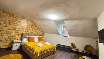 Een slaapkamer met een stenen muur, een bed met een geel dekbed, een raam met jaloezieën en een klein tafeltje met een stoel.