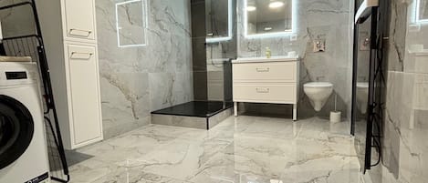 Une salle de bain moderne avec un sol en marbre, un lave-linge, une cabine de douche en verre et un meuble-lavabo avec un miroir.