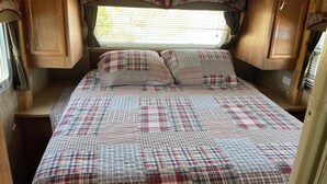 A bedroom with a plaid bedspread, wooden cabinets, and a window with blinds. - RV By The Sea. 3 Rogers Cove Rd. Cobbs Arm, NL. You, the Ocean and Mother Nature (Cobbs Arm)