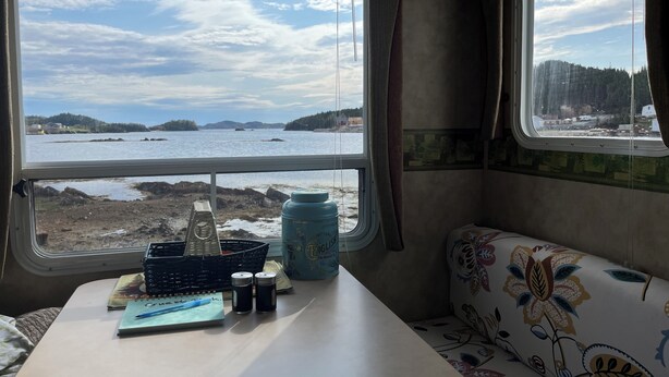 A cabin interior with a table, a notebook, salt shakers, a water jug, and a view of a coastal landscape through large windows. - RV By The Sea. 3 Rogers Cove Rd. Cobbs Arm, NL. You, the Ocean and Mother Nature (Cobbs Arm)