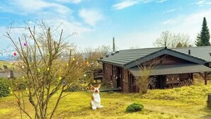 A wooden cabin with a sloped roof, surrounded by a grassy area with a small tree and colorful decorations.