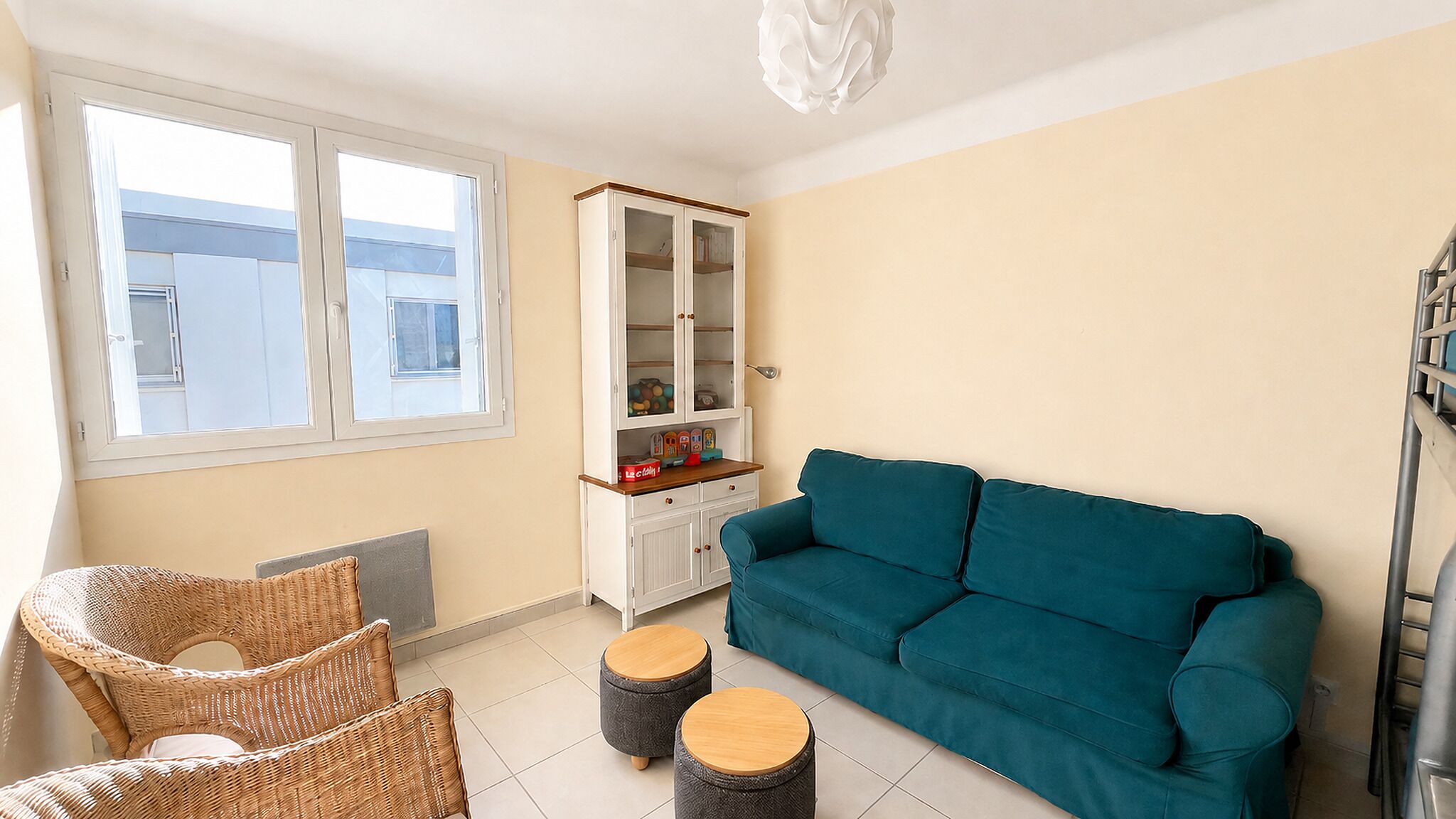 Ein Wohnzimmer mit einem blaugrünen Sofa, Korbstühlen, einer Vitrine und einem Fenster mit Jalousien.