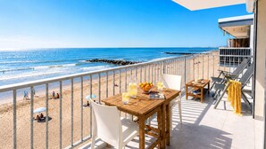 Un balcon avec une table en bois, des chaises blanches et une vue sur la plage et l’océan.