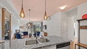 A modern kitchen with granite countertops, stainless steel sinks, and a dishwasher. There are pendant lights, a dining area with blue chairs, and a view of the outdoors. - Hot Tub + Pool | 2 Bed 2 Bath Ground Suite (West Kelowna)