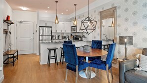 A modern kitchen with white cabinets, a stainless steel refrigerator, and a wooden dining table with blue upholstered chairs. - Hot Tub + Pool | 2 Bed 2 Bath Ground Suite (West Kelowna)