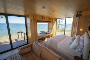 A wooden cabin with a bed, a sofa, and a table by a large window overlooking a body of water. - Wilding Hotels (Quinlan)