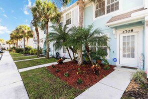 Una hilera de casas urbanas azul claro con un jardín delantero bien cuidado, que presenta palmeras y jardinería con acolchado rojo.
