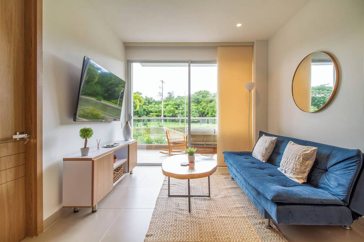 A modern living room with a blue sofa, a flat-screen TV mounted on the wall, a small table with a plant, and a sliding glass door leading to a balcony with a view of greenery.