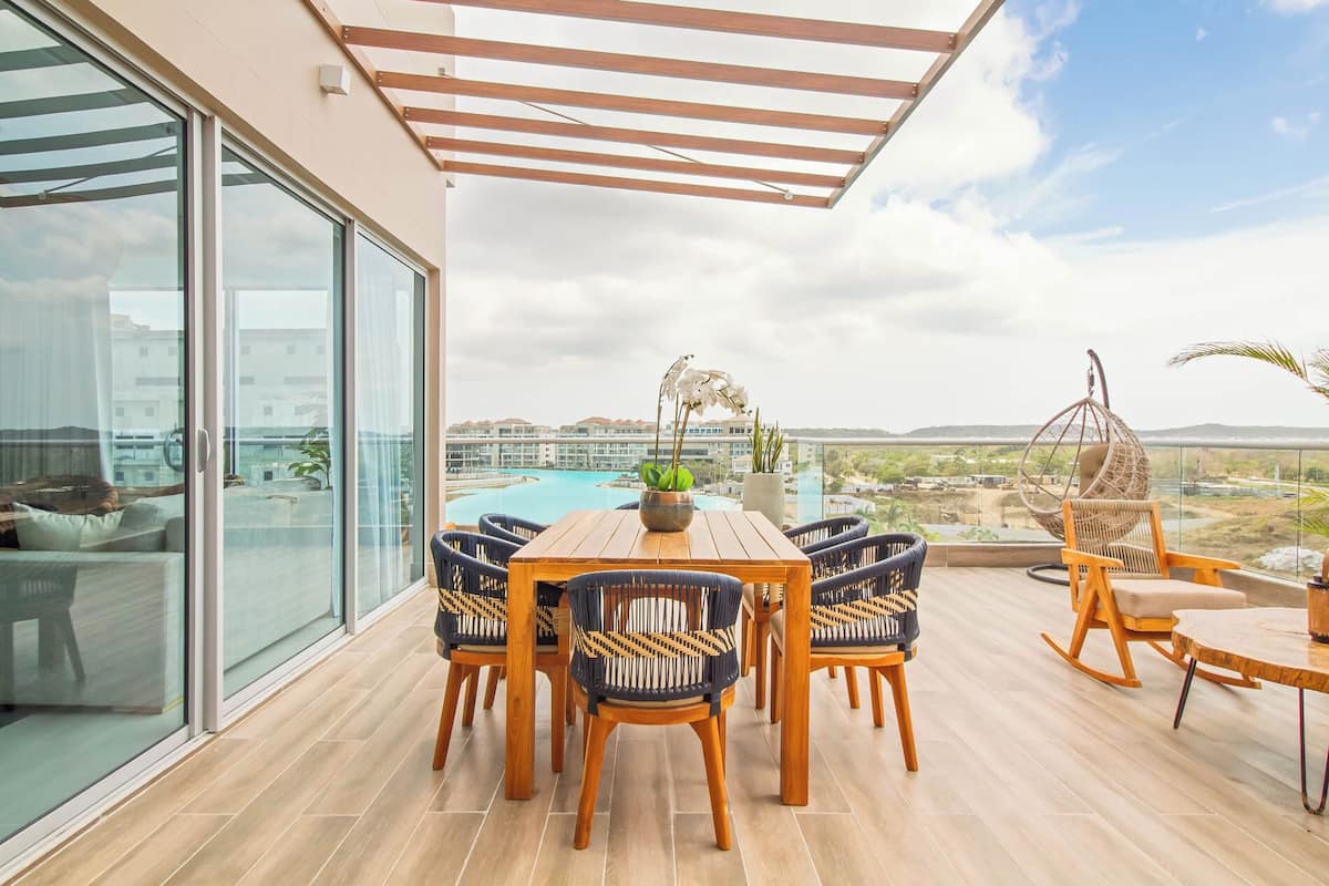 A balcony with a wooden table and chairs, a hanging chair, and a view of a swimming pool and buildings.
