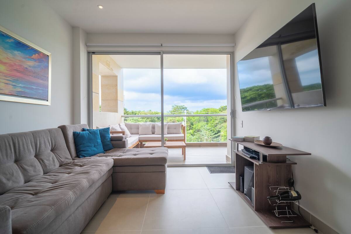 A modern living room with a grey sectional sofa, a wooden coffee table, and a TV mounted on the wall. There is a balcony with a glass door leading to a lush green landscape.