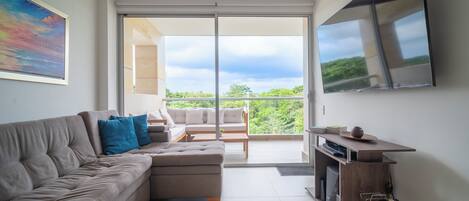 A modern living room with a grey sectional sofa, a wooden coffee table, and a TV mounted on the wall. There is a balcony with a glass door leading to a lush green landscape.
