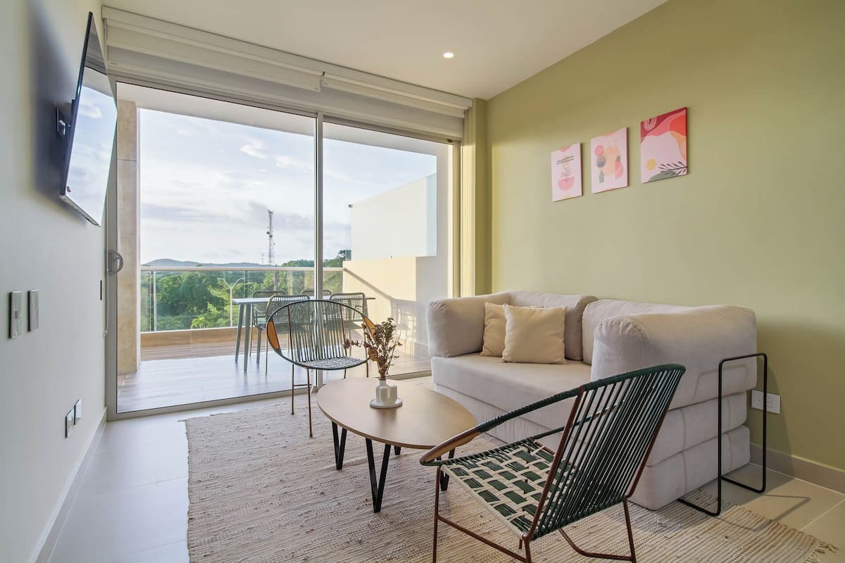 A modern living room with a sofa, a coffee table, and two chairs. There is a sliding glass door leading to a balcony with a view of the city.