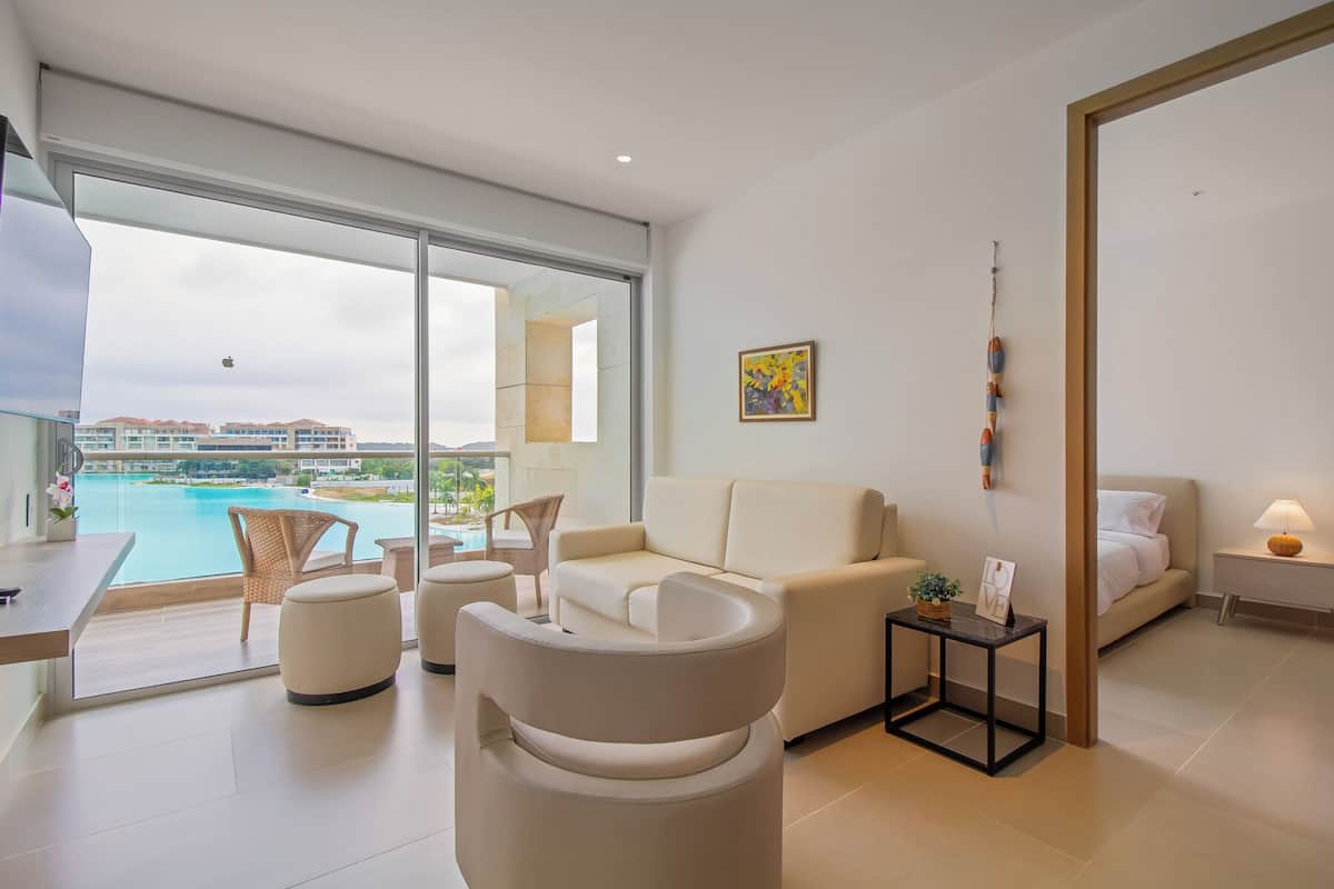 A modern living room with a sofa, armchairs, and a small table. There is a large window with a view of a pool and buildings.