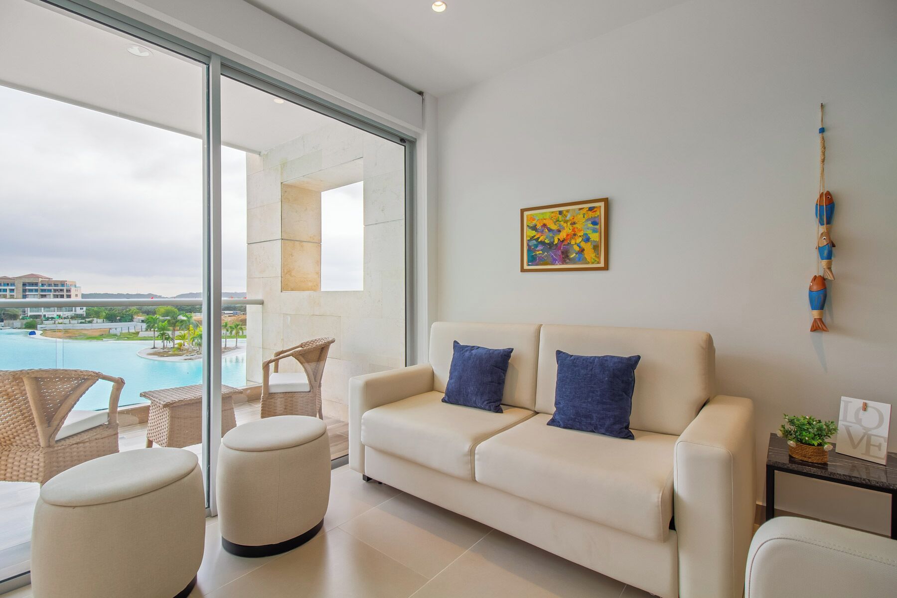 Una sala moderna con un sofá color crema, dos cojines azules, una silla de mimbre, un reposapiés redondo y vistas a una piscina y edificios al fondo.