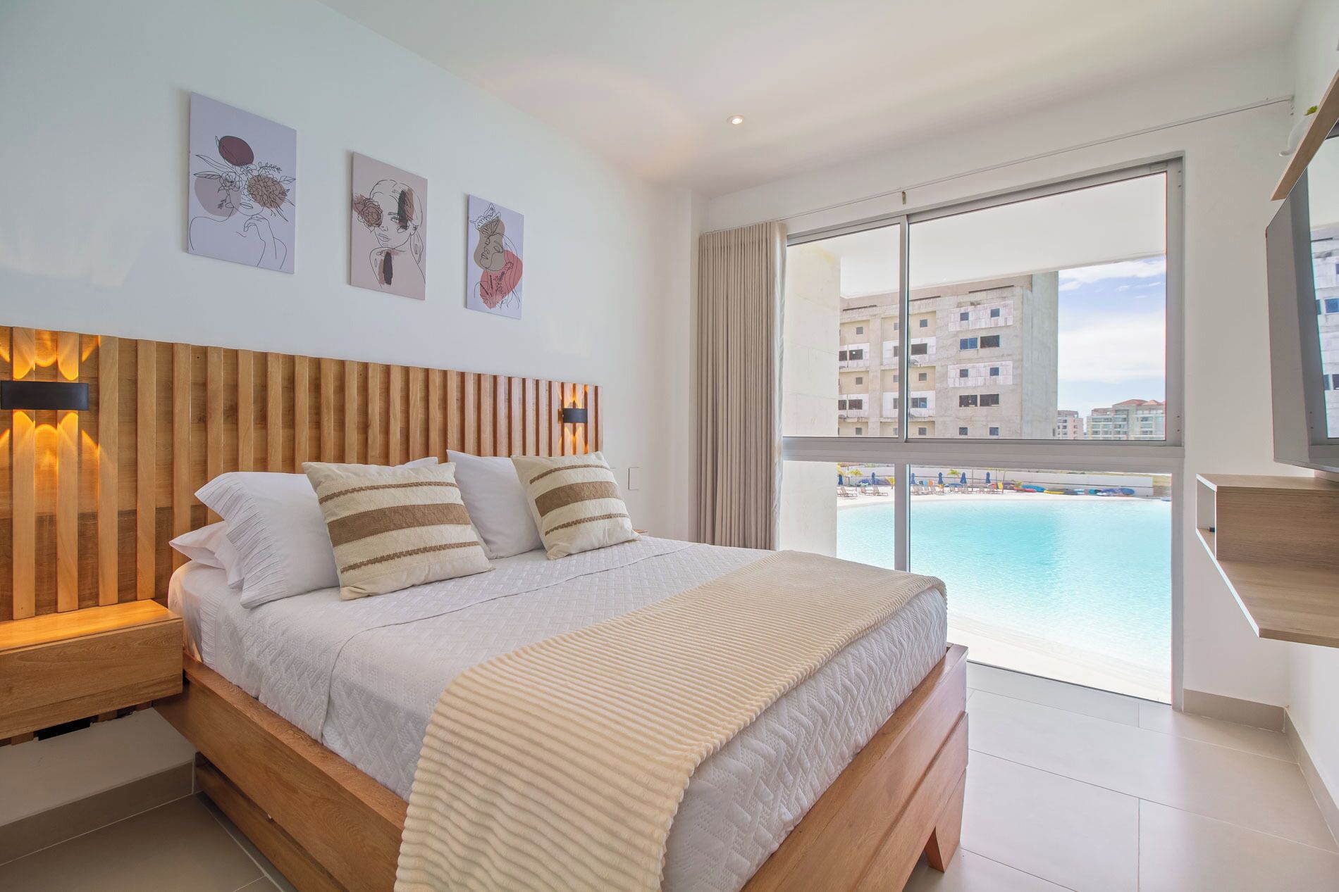 Un dormitorio moderno con cabecera de madera, cama grande con almohadas a rayas y vista a una piscina y edificios a través de ventanales.