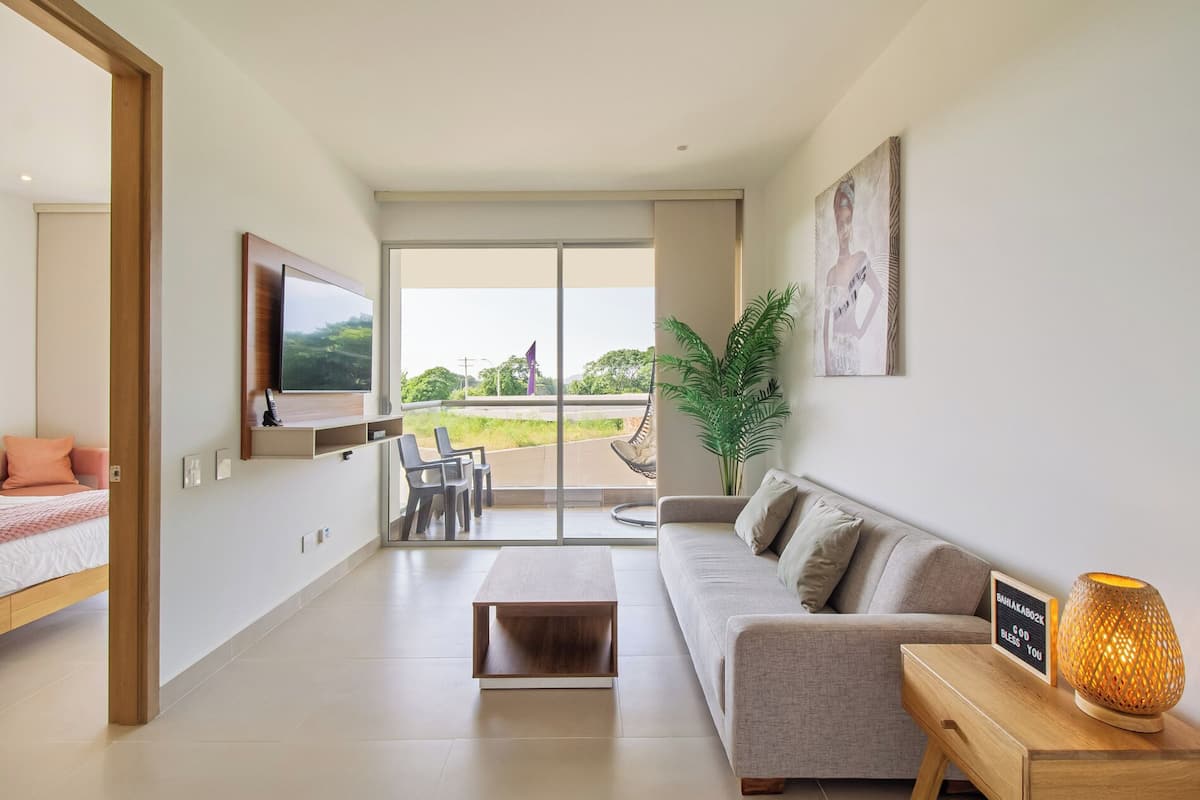 A modern living room with a sofa, a coffee table, a TV, and a large window offering a view of the outdoors.