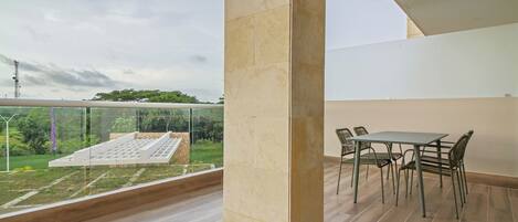 A balcony with a view of greenery and a white structure, a wooden floor, and a table with chairs.