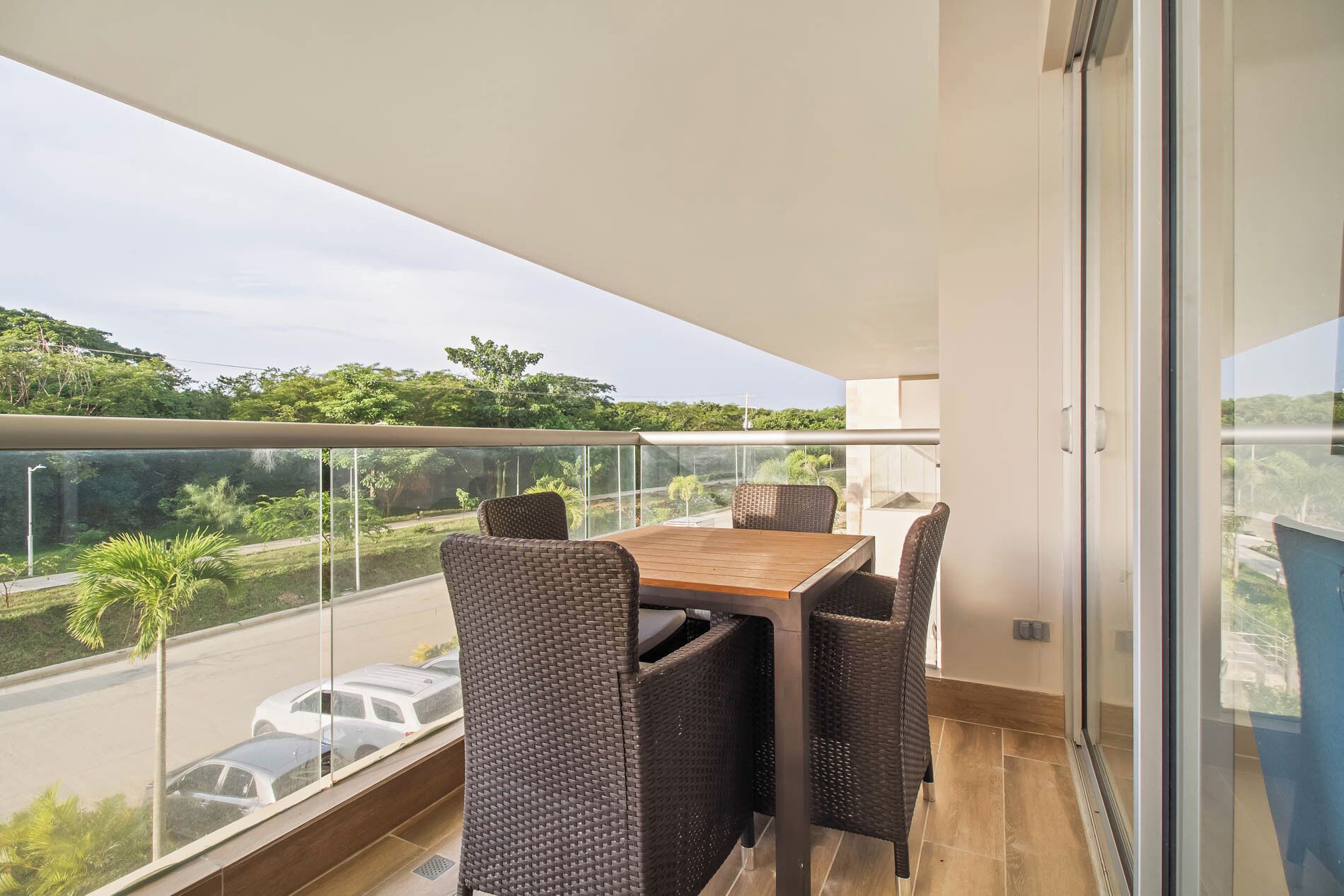 A balcony with a wooden table and wicker chairs, offering a view of a road and greenery.