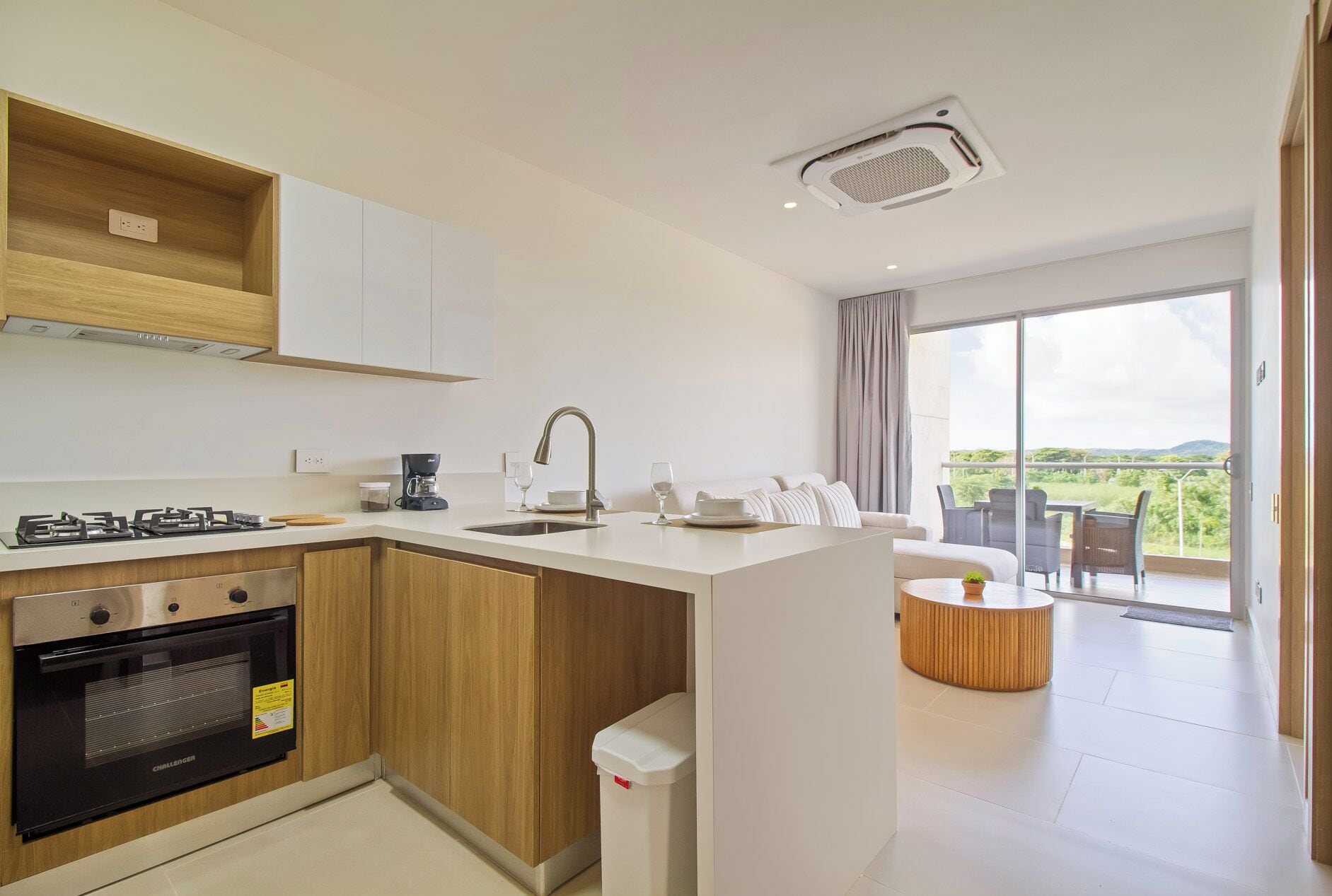 A modern kitchen with a gas stove, oven, and coffee maker. There is a dining area with a table and chairs, and a balcony with a view.