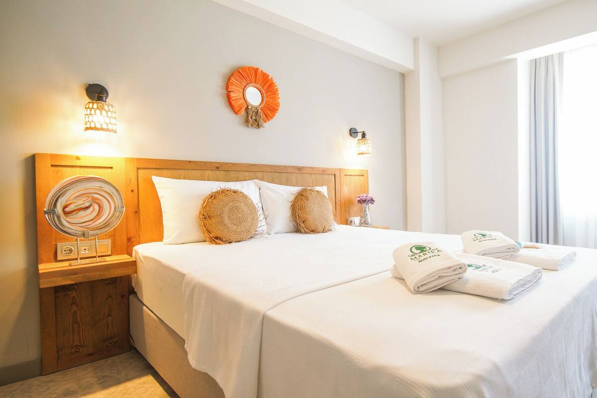 Habitación de hotel con cabecera de madera, cama con ropa blanca, dos lámparas fijadas a la pared, un espejo decorativo y un ventanal con cortinas.