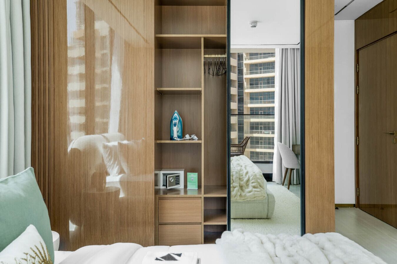 A modern hotel room with a large wardrobe, a bed with white bedding, a small seating area with a chair, and a view of the city through the window.