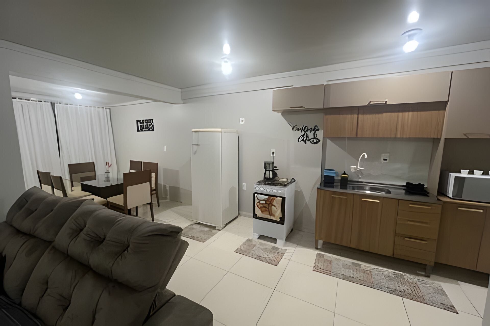 A modern living room with a sofa, dining table, and chairs, a kitchen with wooden cabinets and a stove, and a refrigerator.