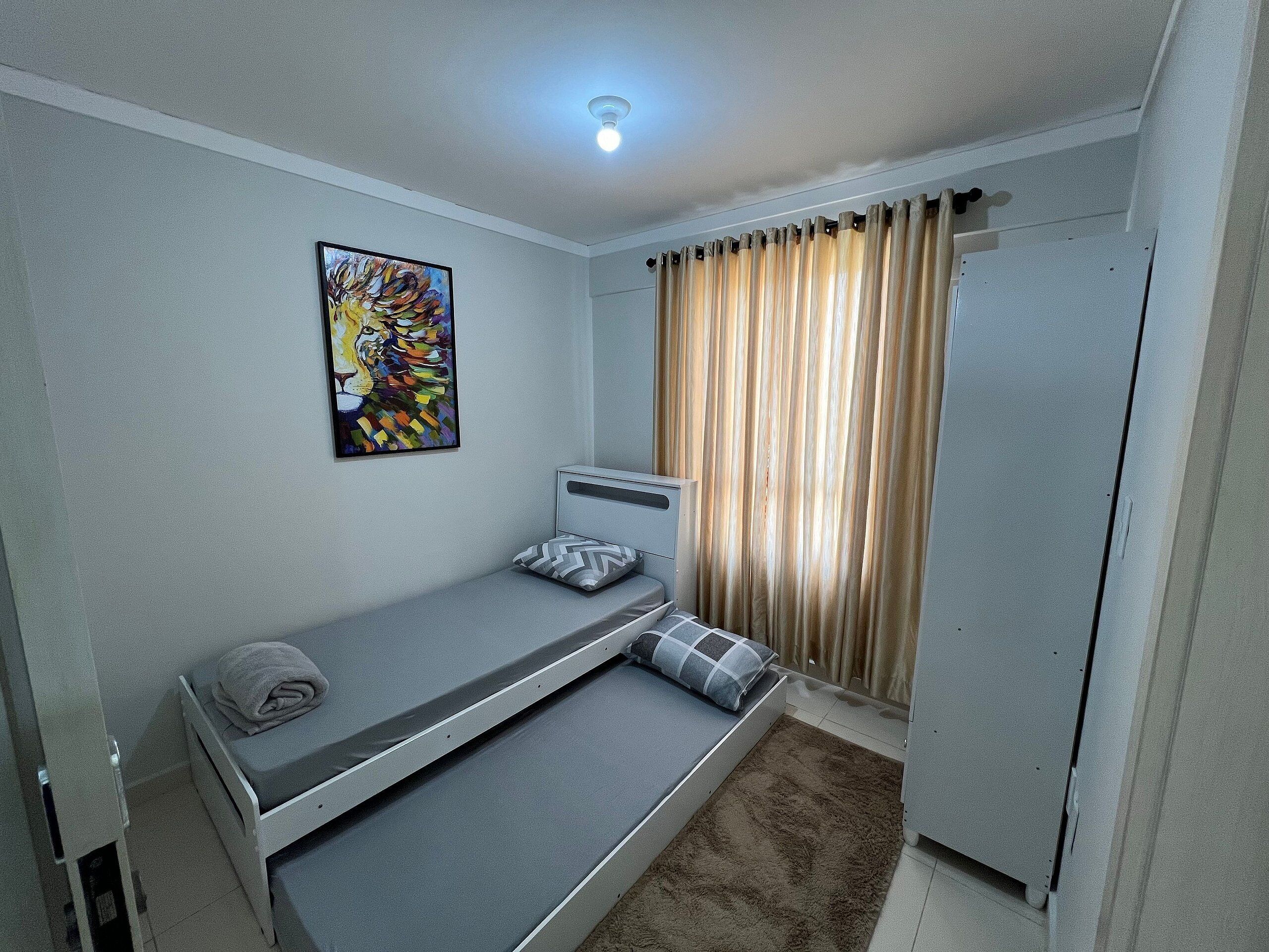 A small, single-bed room with a gray mattress, a gray pillow, a beige rug, a ceiling light, a window with beige curtains, and a colorful abstract painting on the wall.