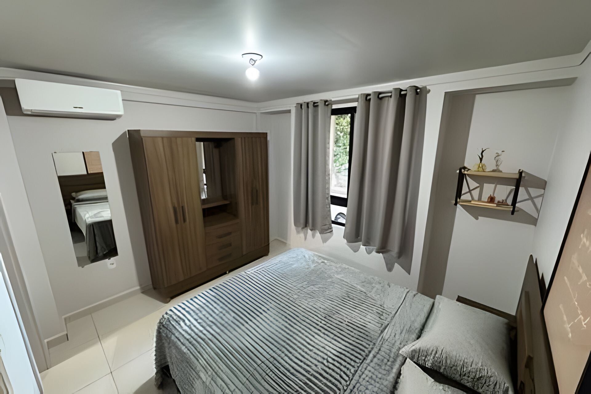 Un dormitorio con cama, armario, unidad de aire acondicionado integrada, ventana con cortinas y un pequeño estante con adornos.