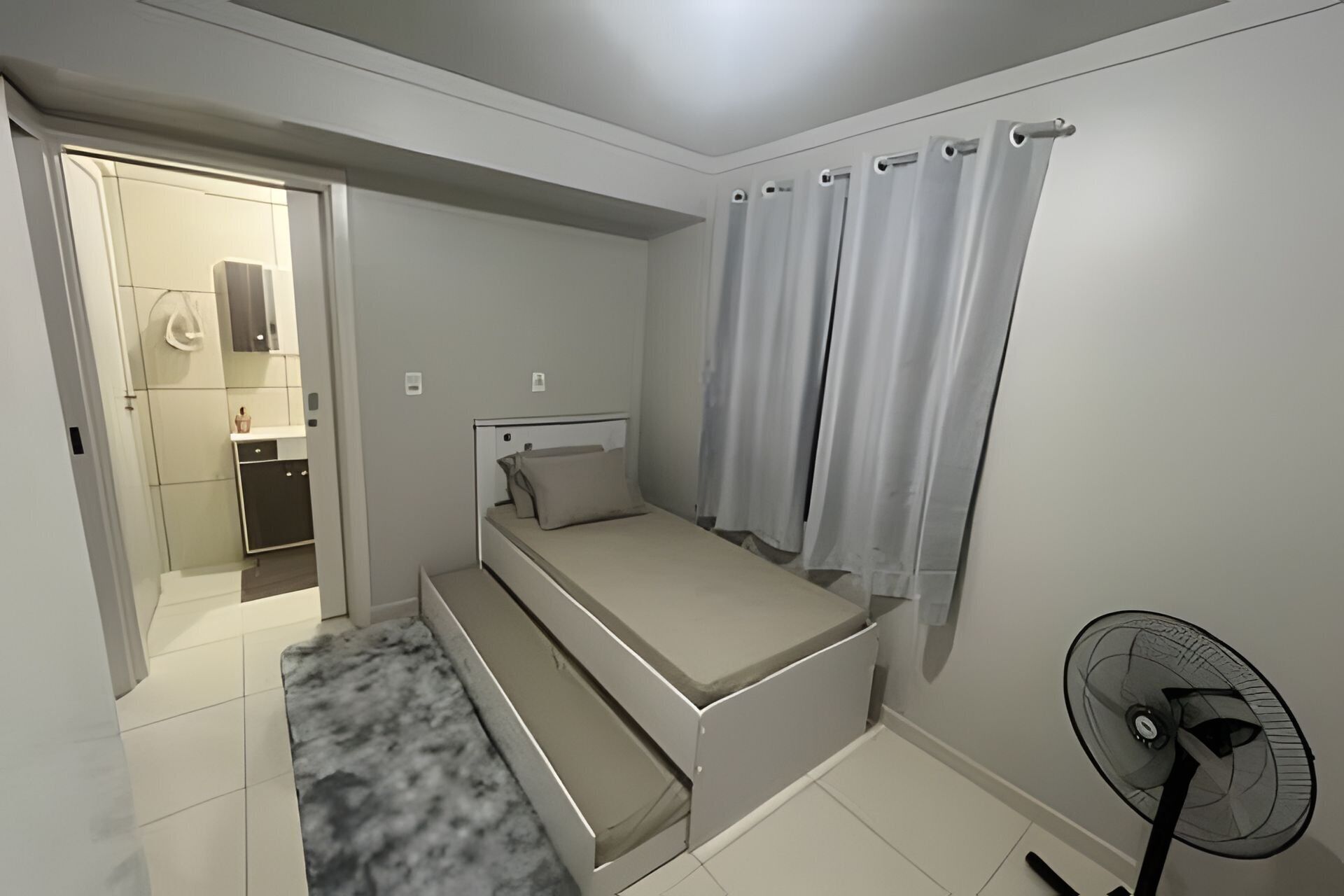 Une chambre à coucher avec un lit, une table de chevet, un ventilateur et une salle de bain visible à travers une porte ouverte.