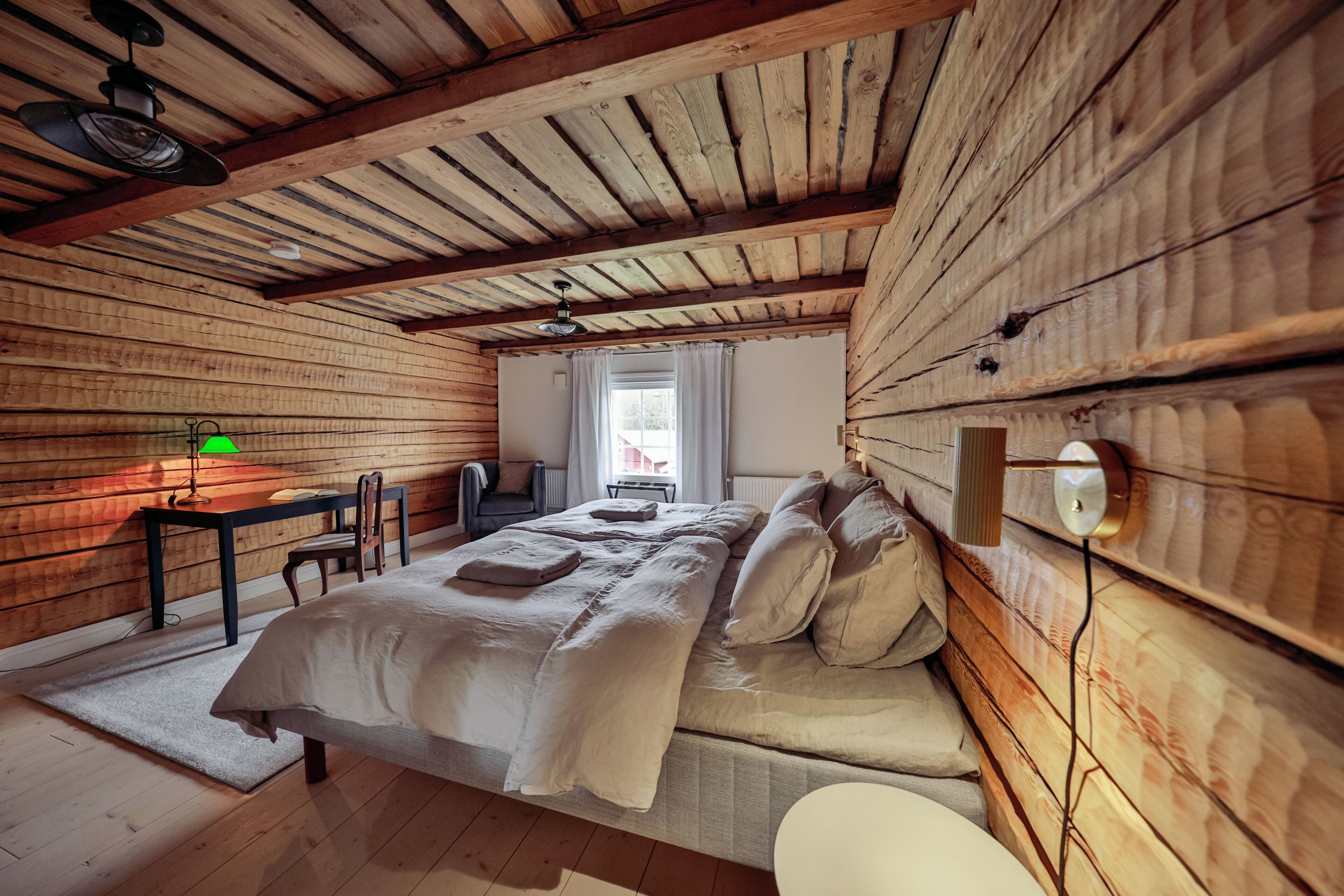 A cozy bedroom with a wooden ceiling, a bed with white linens, a desk with a lamp, and a chair.