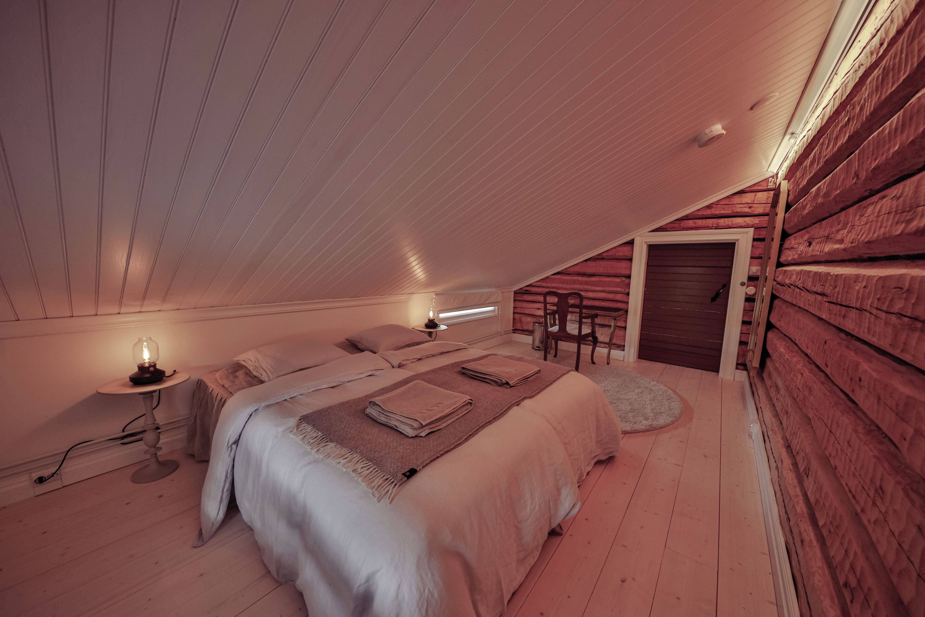 Une chambre confortable située sous les toits, avec un plafond en pente, un lit recouvert d’une couette blanche, une table de chevet avec une lampe et une chaise près de la porte.