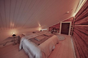 A cozy attic bedroom with a sloped ceiling, a bed with a white comforter, a bedside table with a lamp, and a chair by the door.