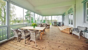 Eine überdachte Terrasse mit Korbmöbeln und einem Holztisch, großen Fenstern und Blick ins Freie.