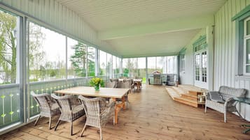 A covered patio with wicker furniture and a wooden table, large windows, and a view of the outdoors.