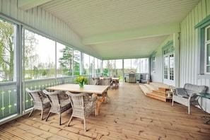 Eine überdachte Terrasse mit Korbmöbeln und einem Holztisch, großen Fenstern und Blick ins Freie.