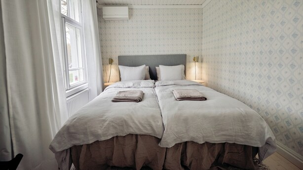 Ein Doppelbett mit grauem Kopfteil, zwei Nachttische mit Lampen und ein Fenster mit Vorhängen.