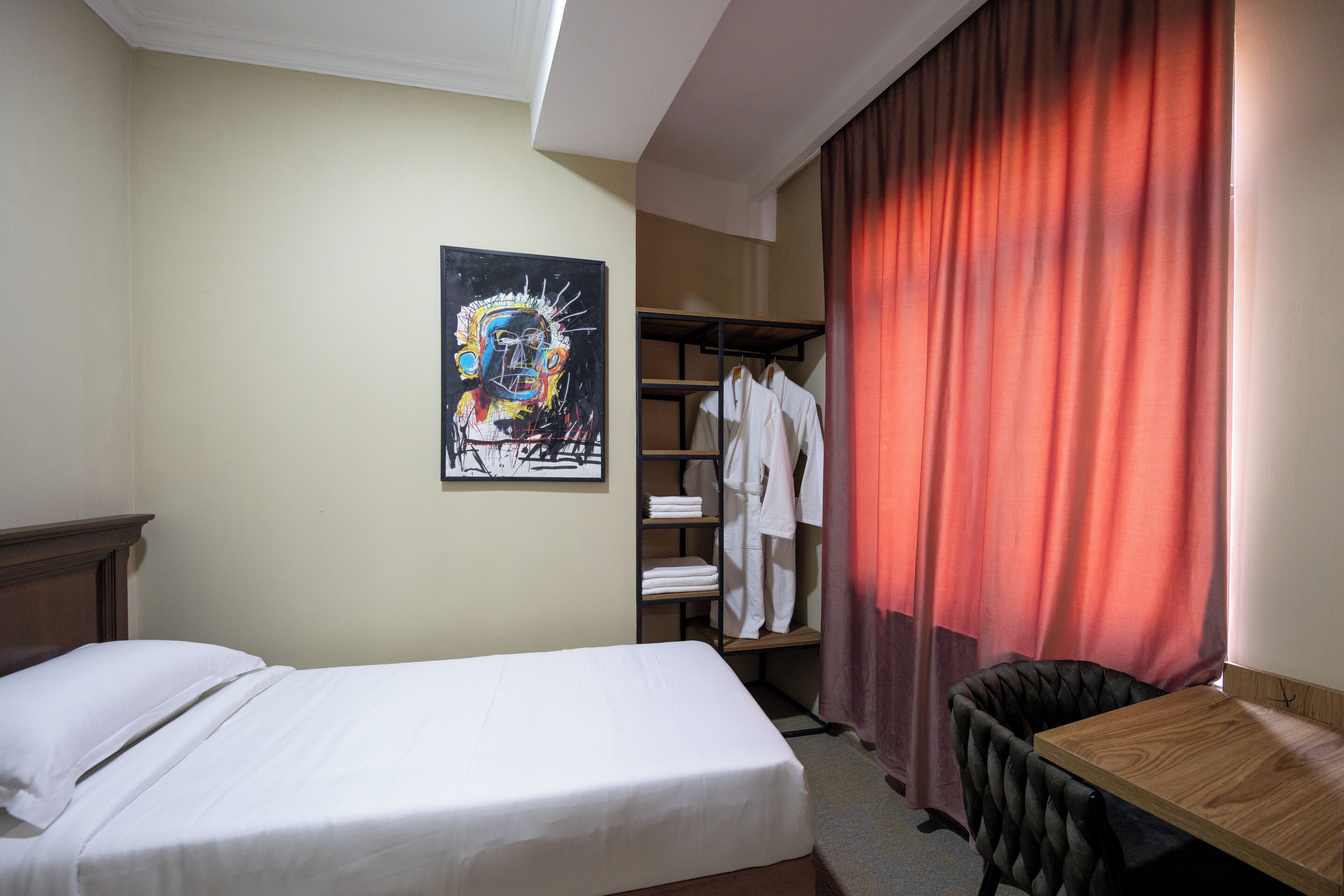 A hotel room with a bed, a wooden desk, a wardrobe, and a painting on the wall.