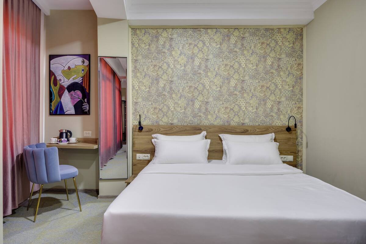 A hotel room with a large bed, a desk with a chair, a painting on the wall, and a patterned wallpaper.