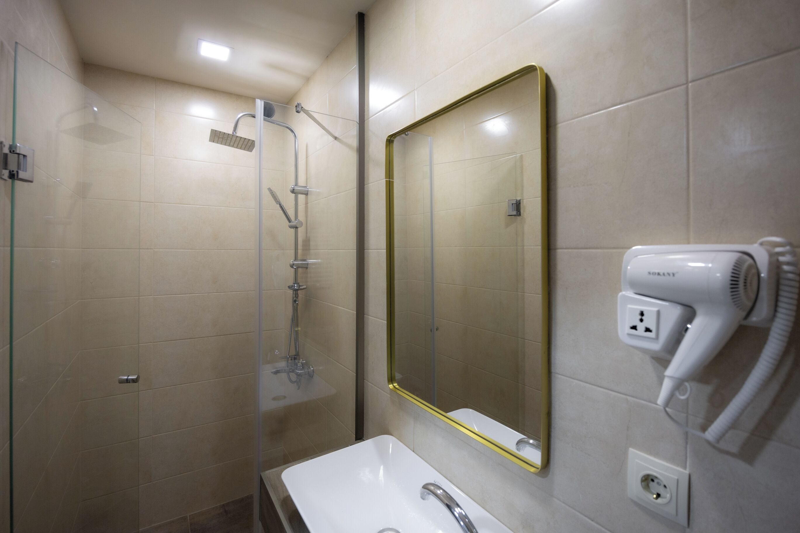 Un baño con ducha envuelta en vidrio, un lavamanos blanco, un espejo con marco dorado y un secador de pelo enchufado.