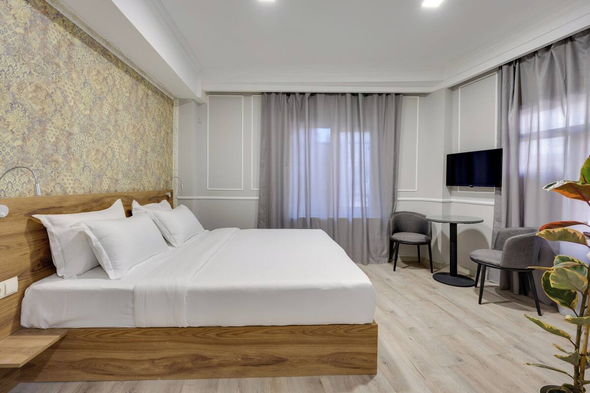A hotel room with a wooden headboard bed, a small round table, two chairs, a TV, and a large window with curtains.