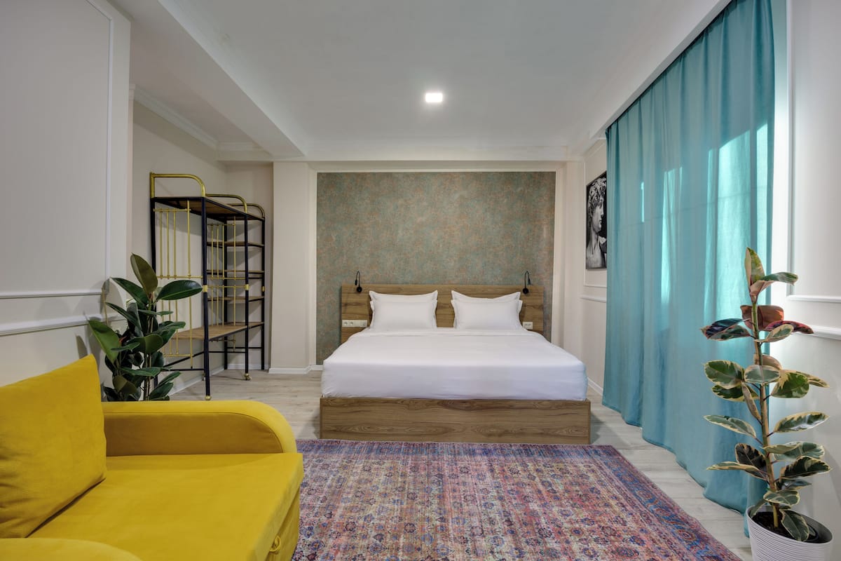 A bedroom with a bed, a yellow sofa, a patterned rug, a plant, and a staircase with a metal railing.