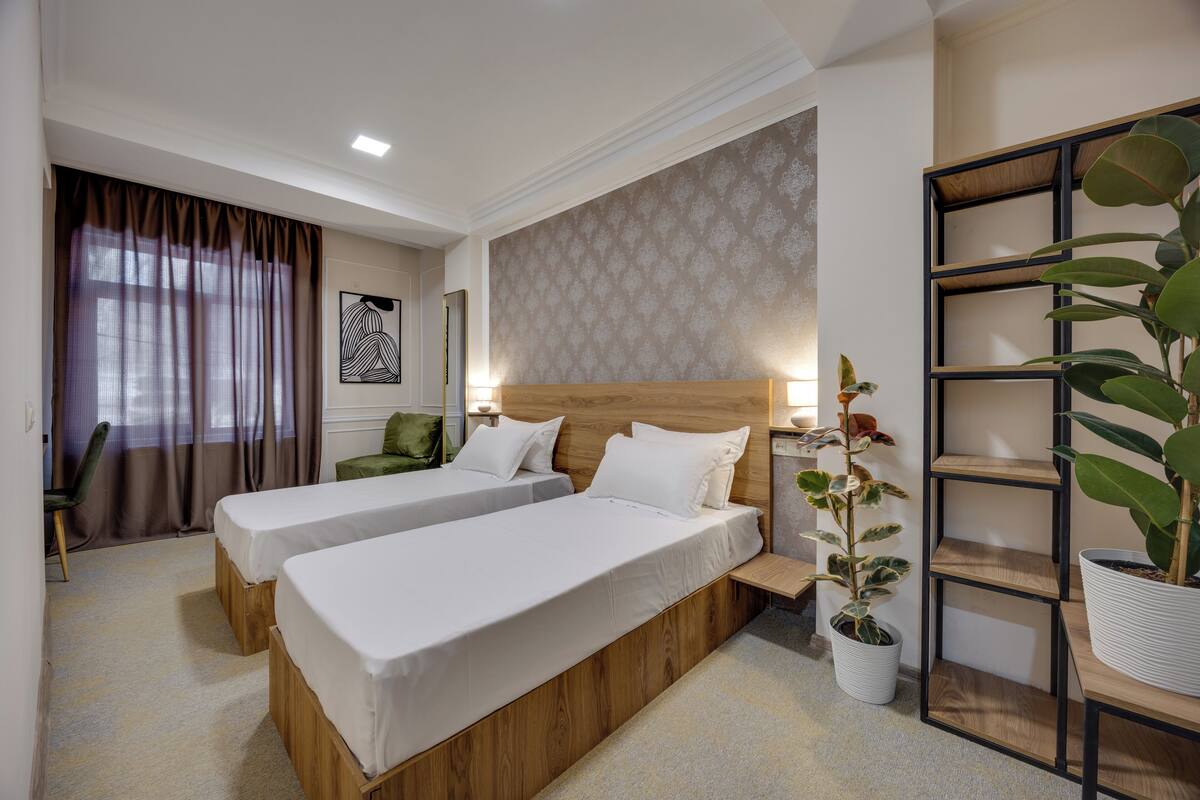 A hotel room with two single beds, a wooden headboard, a potted plant, a bookshelf, and a desk with a chair.
