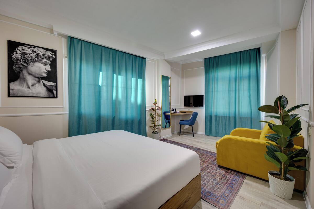 A hotel room with a bed, a desk, a chair, a TV, and a plant.