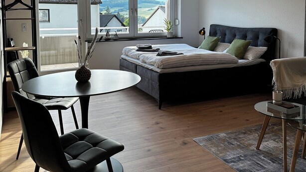 Quarto moderno com cama, mesa de jantar e cadeiras. Há uma janela com vista para casas e colinas.