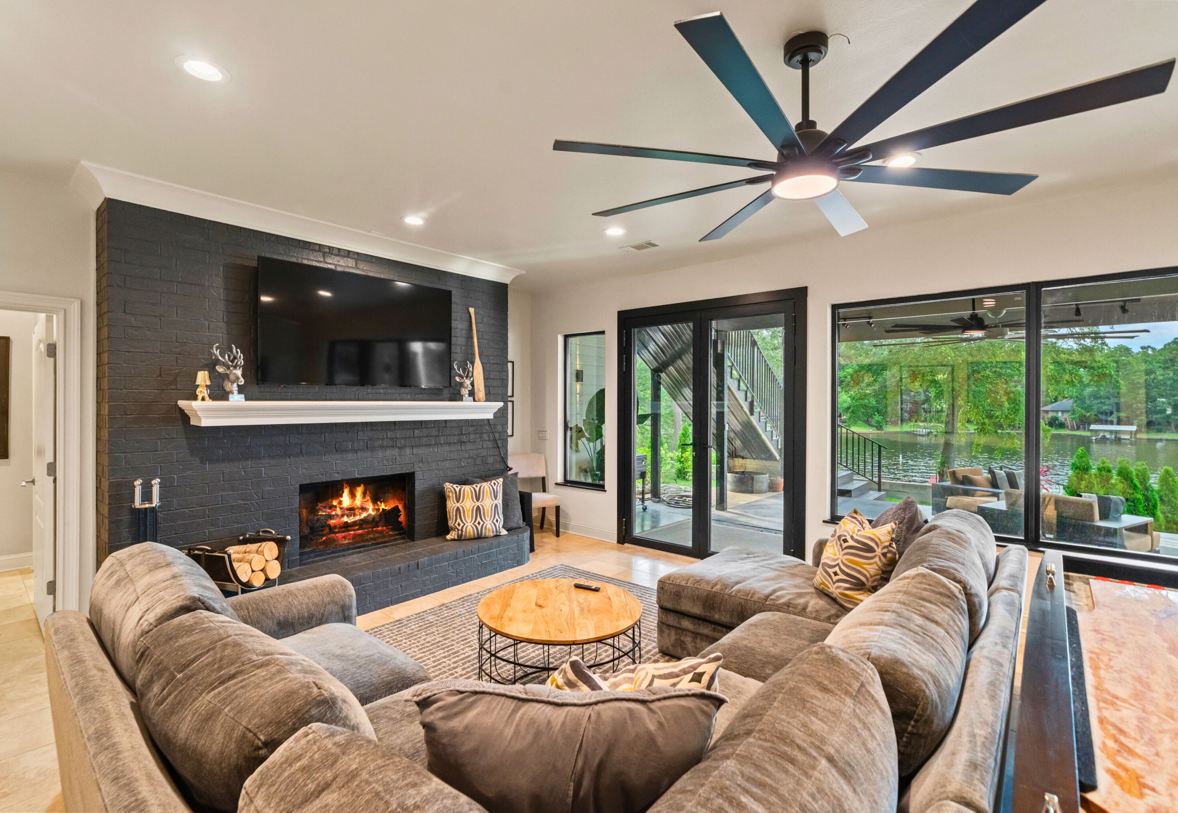 Un salon moderne doté d’une cheminée, d’un ventilateur de plafond et d’une grande fenêtre donnant sur un jardin.