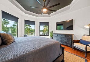 A bedroom with a large bed, a ceiling fan, a TV, and a view of the outdoors.