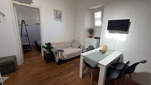 A compact living space with a sofa, a dining table with chairs, a TV mounted on the wall, and a small potted plant.