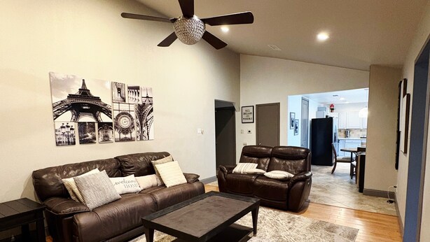 A modern living room with a brown leather sofa, a matching armchair, a coffee table, and a large rug. There is a ceiling fan, wall art depicting the Eiffel Tower, and a door leading to another room.