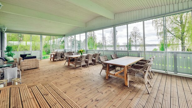 A covered outdoor patio with wooden flooring, wicker furniture, and large windows offering views of a lake and trees. - In the eastern wing, 6 people accommodate in a three two-person rooms (Unknown)
